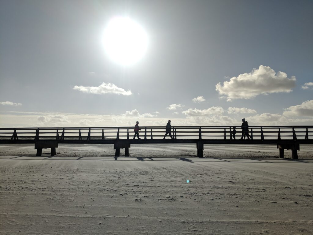 Sonnenuntergang über der Seebrücke in Sankt Peter Ording mit schattenhaften Spaziergängern und weitem Sandstrand im Herbst.