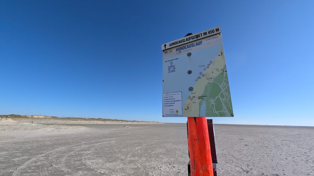 Wegweiser auf einem roten Holzpfahl am Strand von St. Peter-Ording mit Umgebungskarte zum Hundeauslaufgebiet in 450 Metern Entfernung.