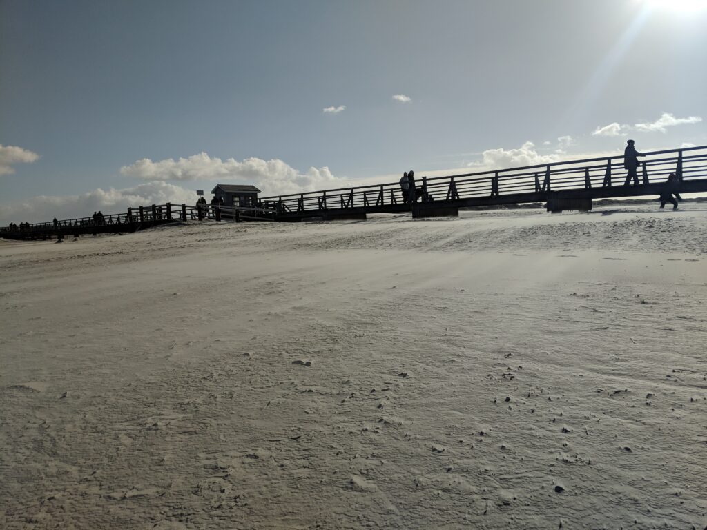 Ein hölzerner Badesteg führt über den weiten Sandstrand von St. Peter-Ording bei hellem, strahlendem Sonnenlicht.