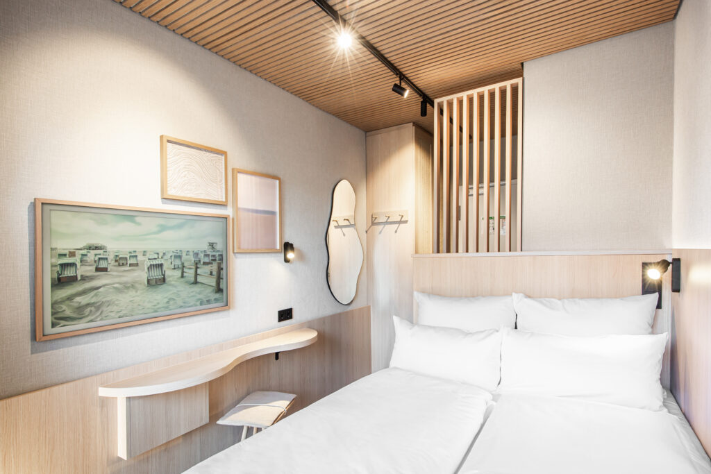 Innenansicht der neu gestalteten Mini Bude in der Bude54 mit Boxspringbett, Eichenholz-Elementen und Samsung The Frame TV.